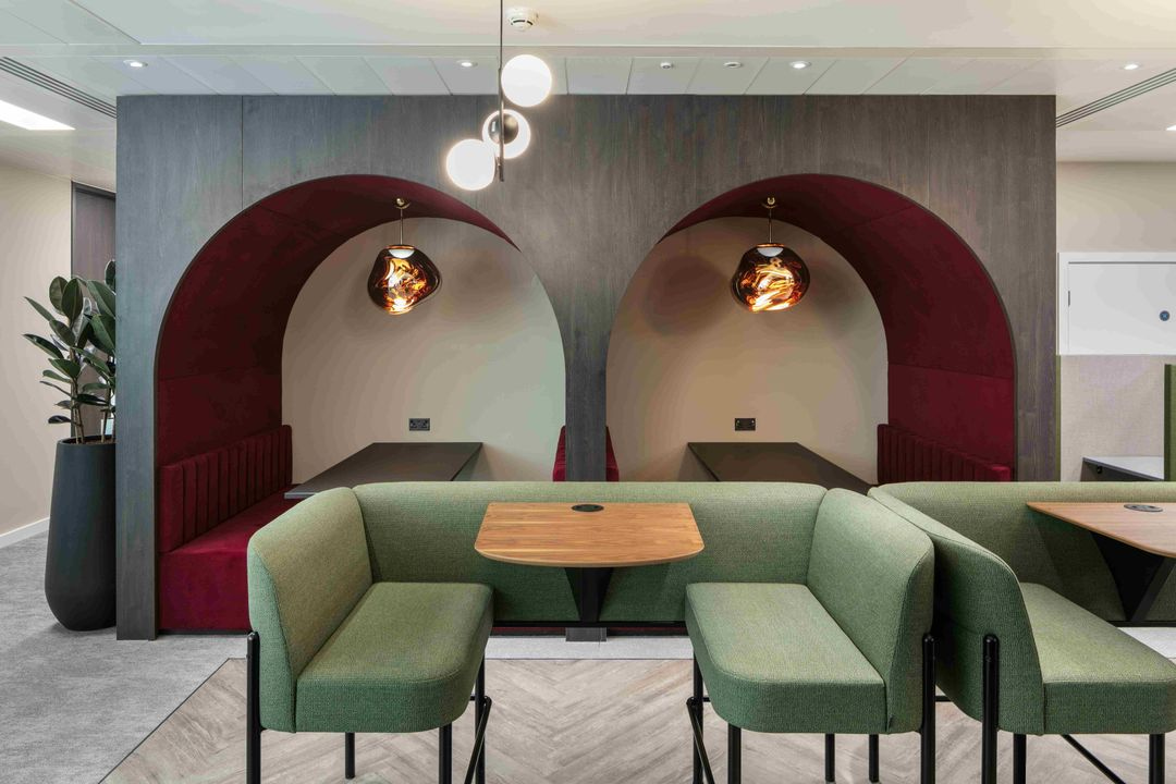 Modern office breakout space featuring arched booth seating with burgundy upholstery, stylish pendant lighting, and green collaborative sofas with integrated tables.