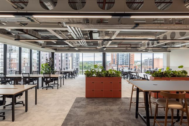 Spacious modern office space with floor-to-ceiling windows, exposed industrial ceiling, collaborative desks, and indoor plants overlooking Manchester city centre.