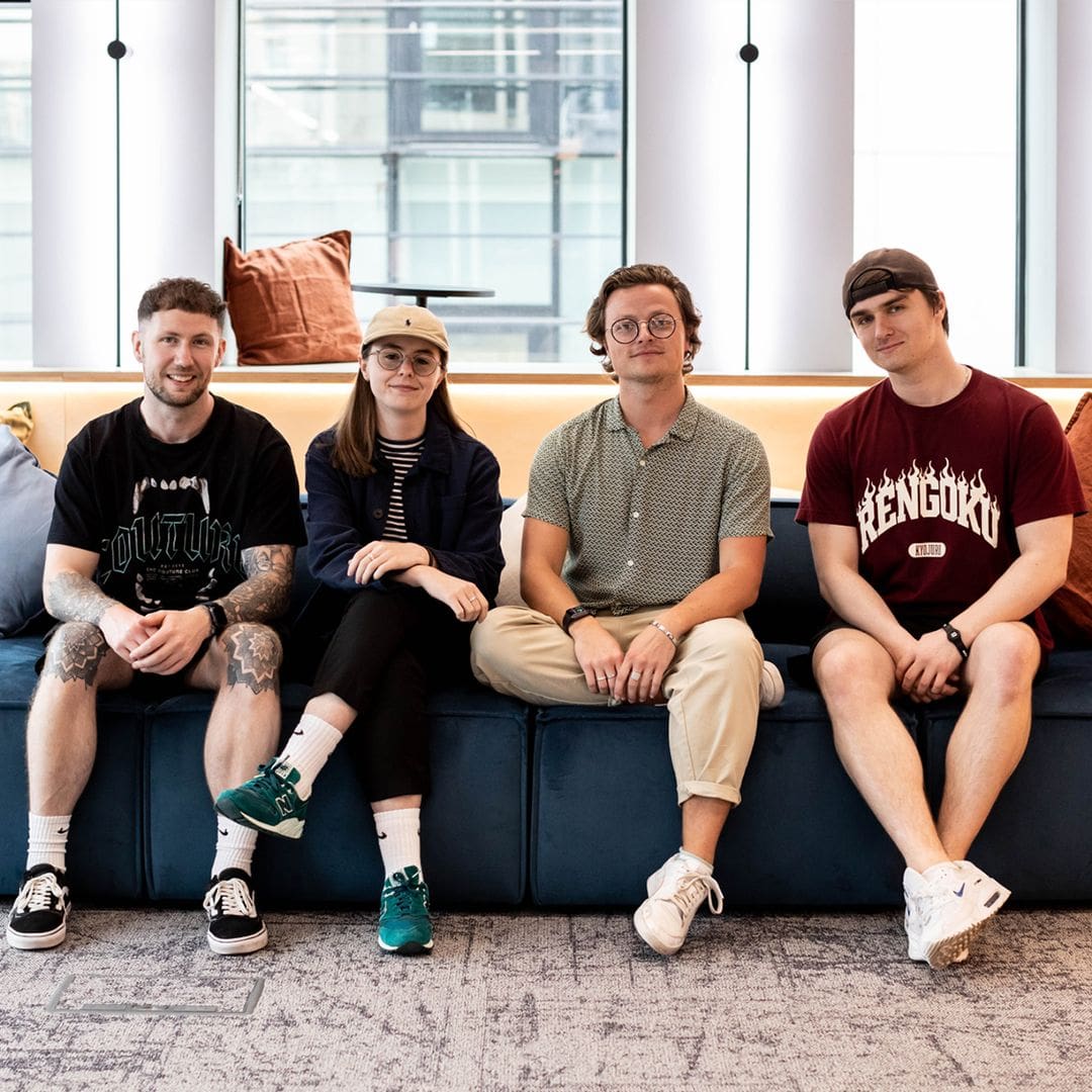 Group of four young professionals sitting on a modern blue sofa in a bright contemporary office lounge with large windows and natural light.
