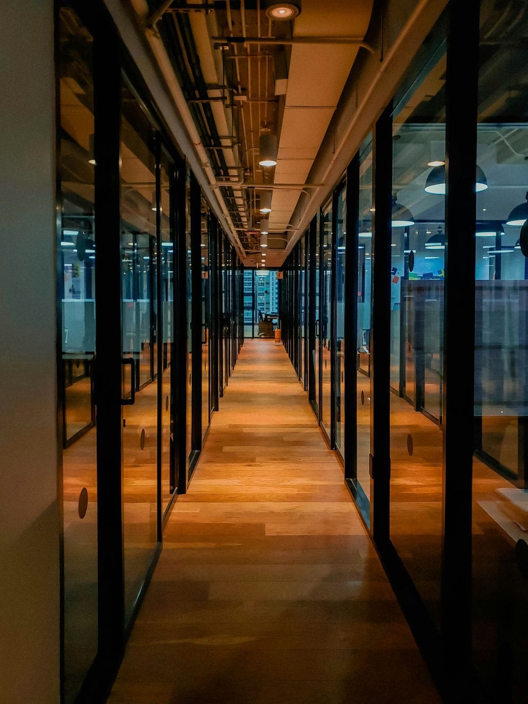 “Contemporary coworking hallway lined with glass private offices and overhead industrial lighting.