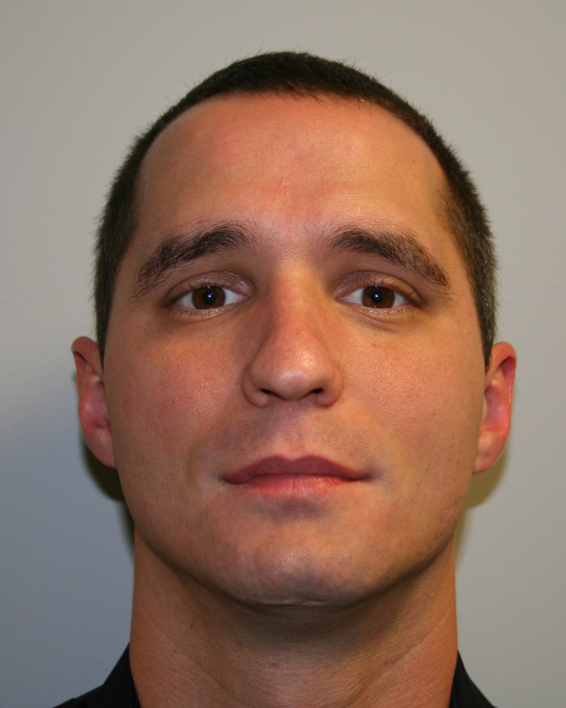 Close-up of a man with short dark hair, brown eyes, and a neutral expression against a plain background.