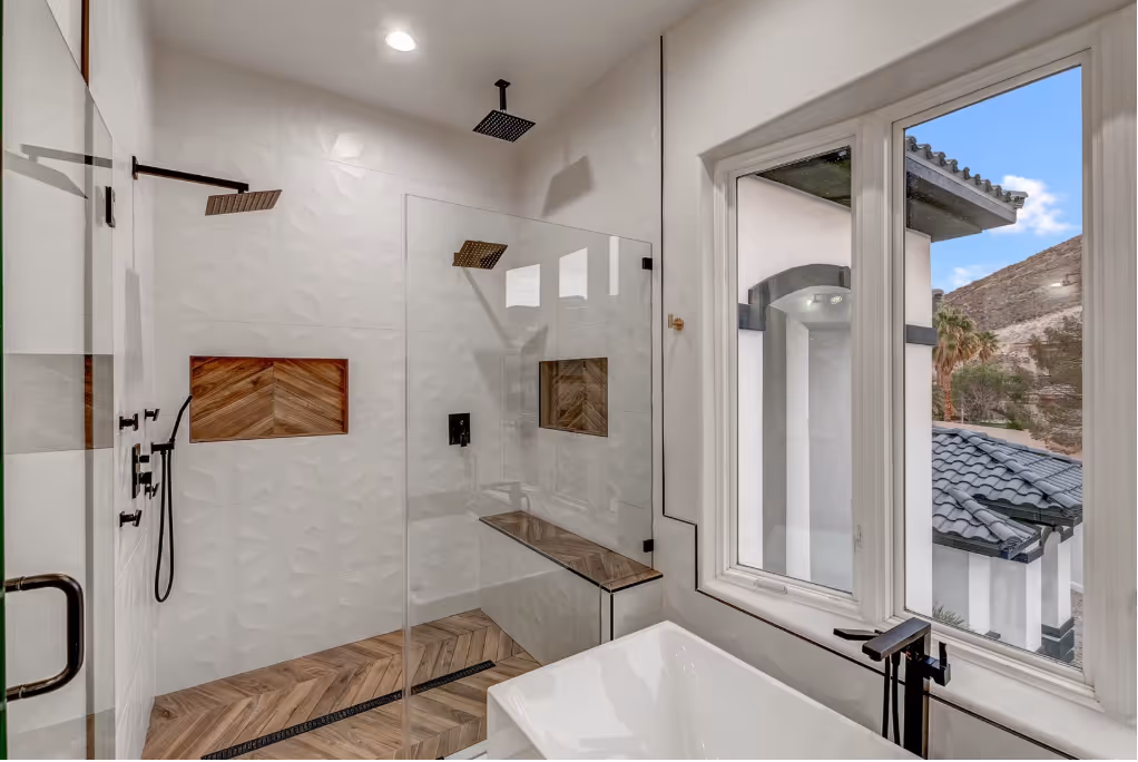 Luxury bathroom with frameless glass shower, wood-accented tile, rainfall showerheads, soaking tub, and mountain view through large windows.