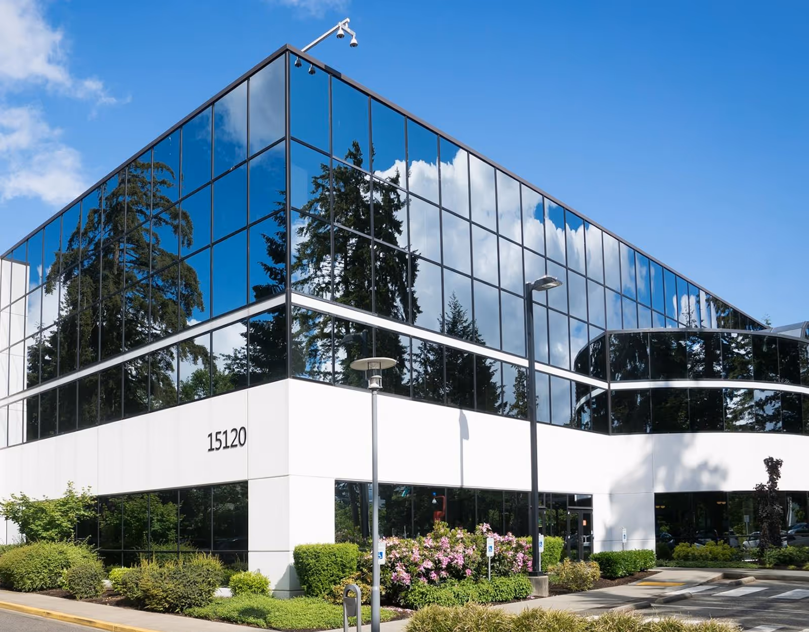 Modern two-story office building with glass windows reflecting trees and sky, surrounded by landscaping and parking.