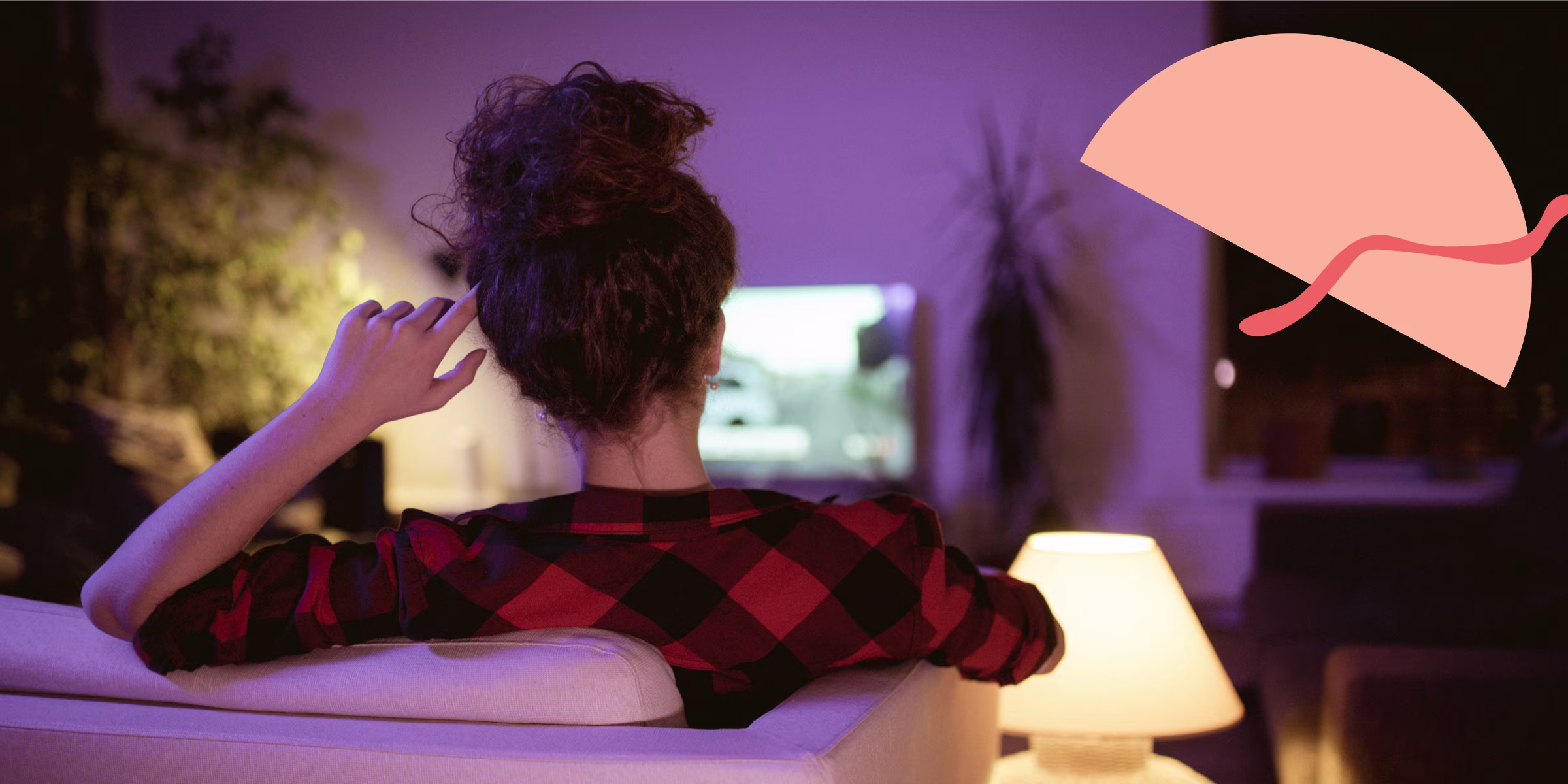 Woman watching TV while sitting on the couch and wearing a plaid shirt, facing away from the camera.