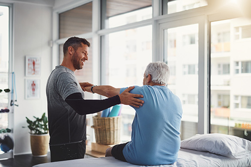 Physical therapist guiding an older man through a shoulder stretch in a rehab clinic