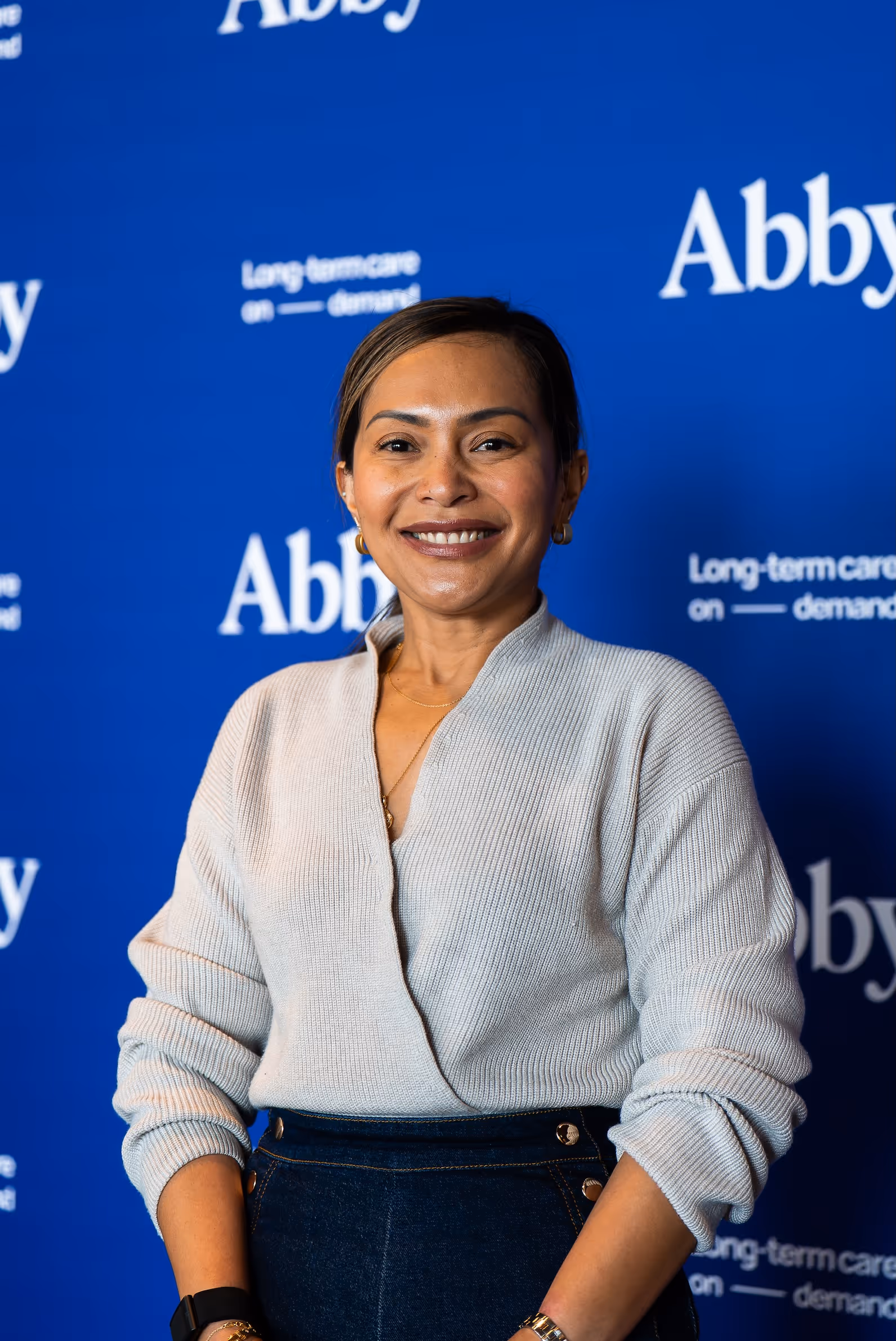 Smiling woman in a light gray sweater and dark pants standing in front of a blue backdrop with repeated 'Abby' and 'Long-term care on demand' text.