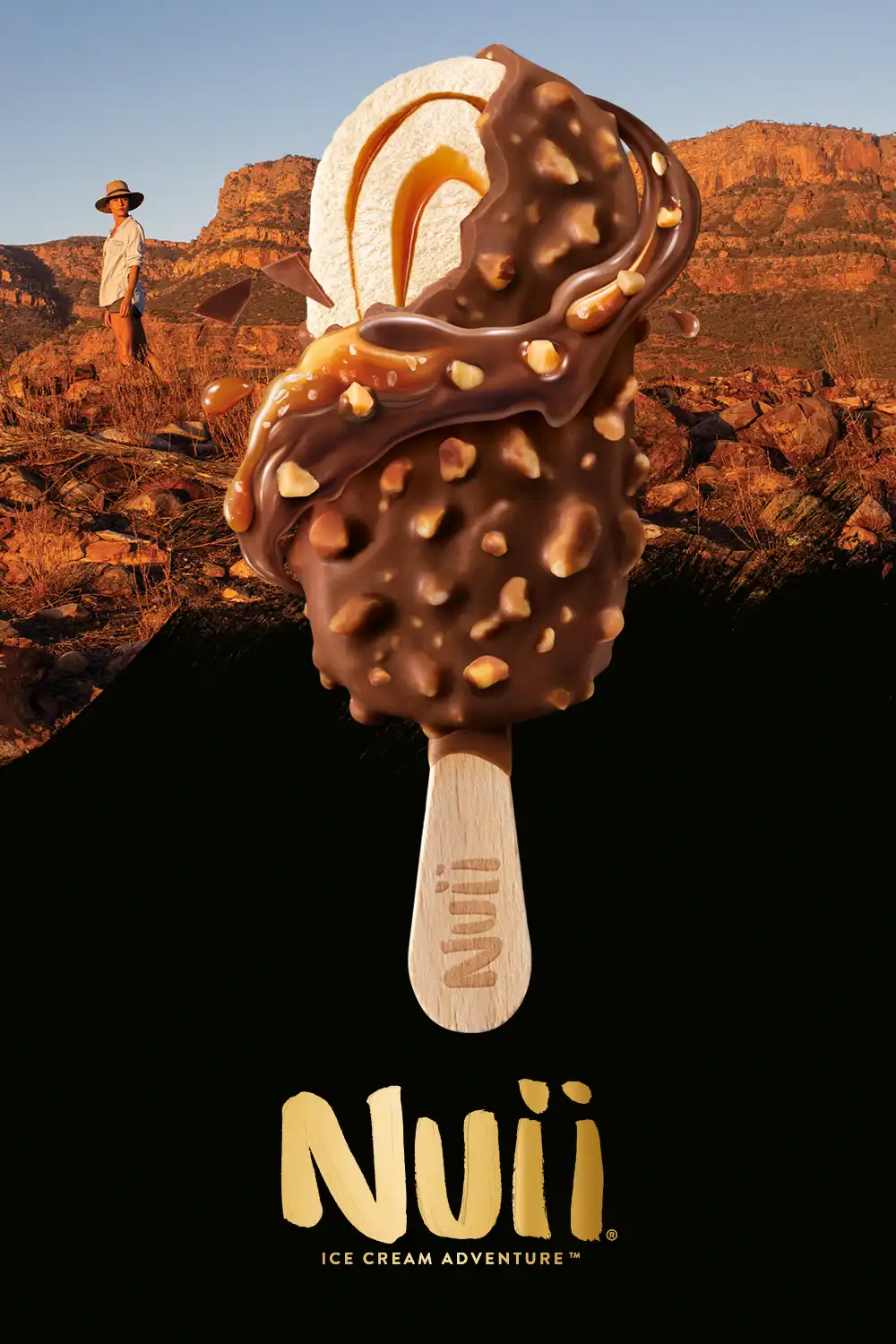 Ice cream bar coated in chocolate and nuts with caramel swirls, set against a rocky desert landscape with a person in a hat in the background, and the text 'Nuii Ice Cream Adventure' below.