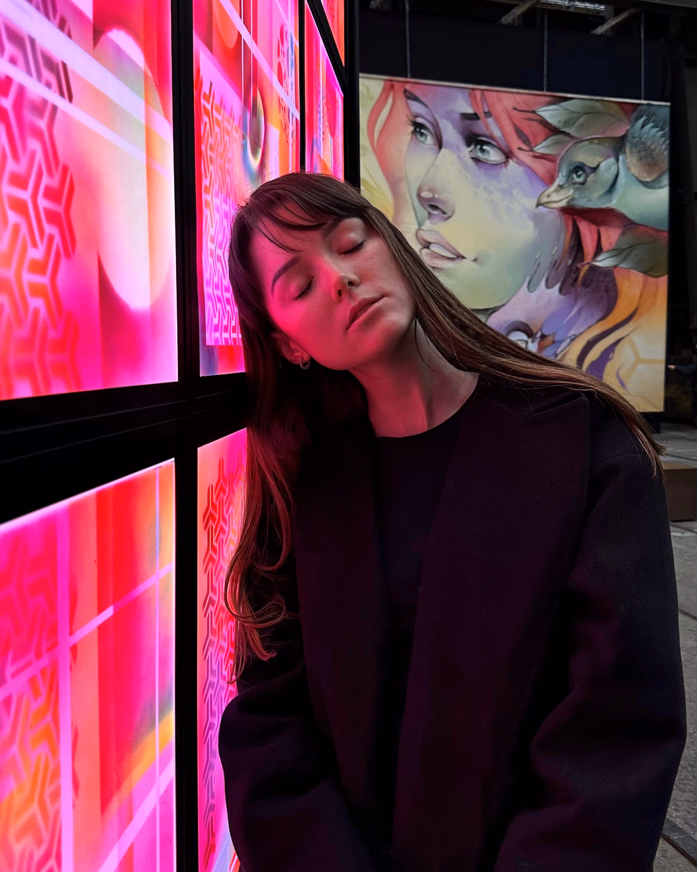 Woman with long brown hair and closed eyes leaning against a brightly lit pink and red patterned illuminated panel, with a colorful mural of a woman and bird in the background.