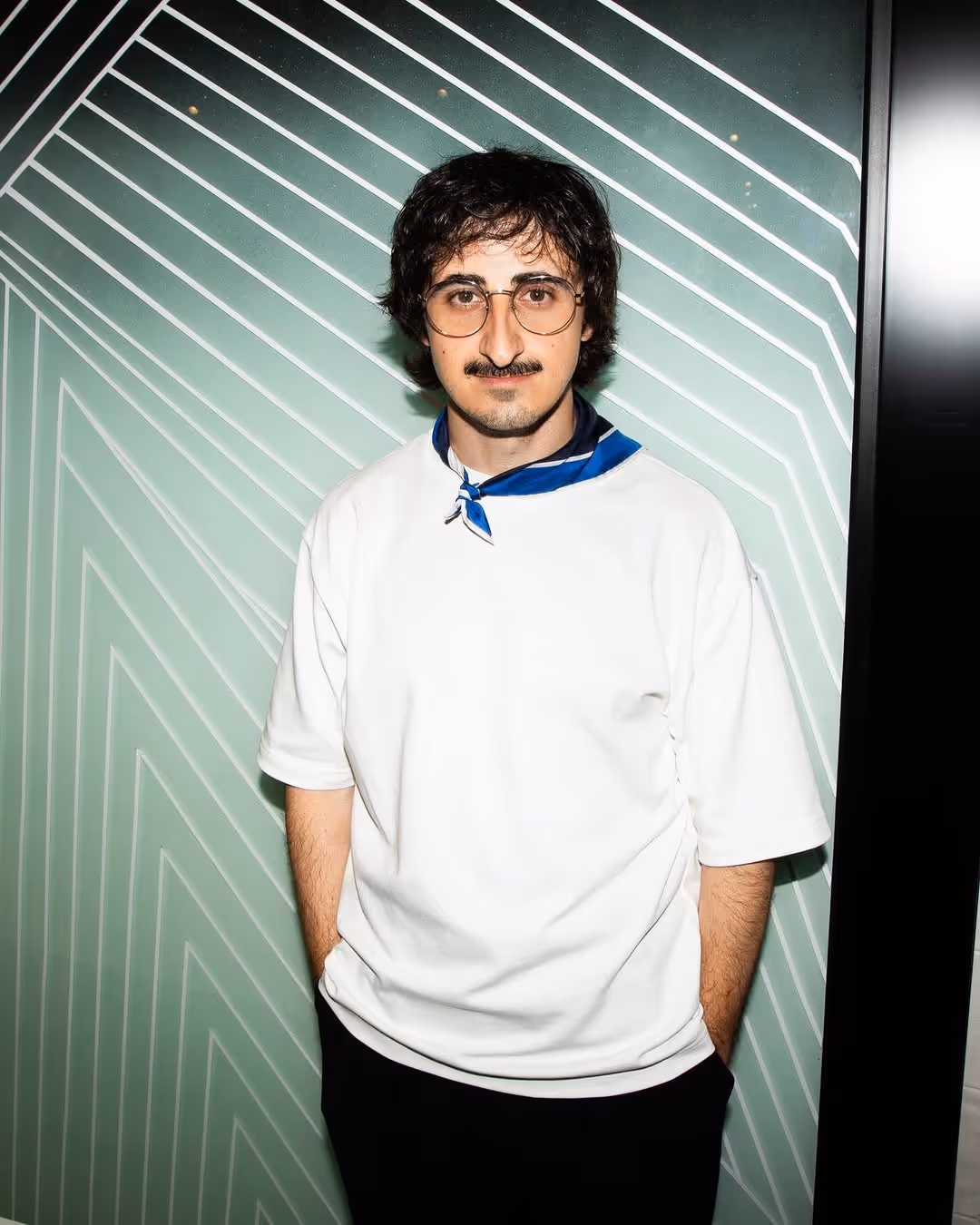 Man with dark hair, glasses, mustache, wearing a white t-shirt and blue neckerchief standing against a light green wall with white geometric lines.