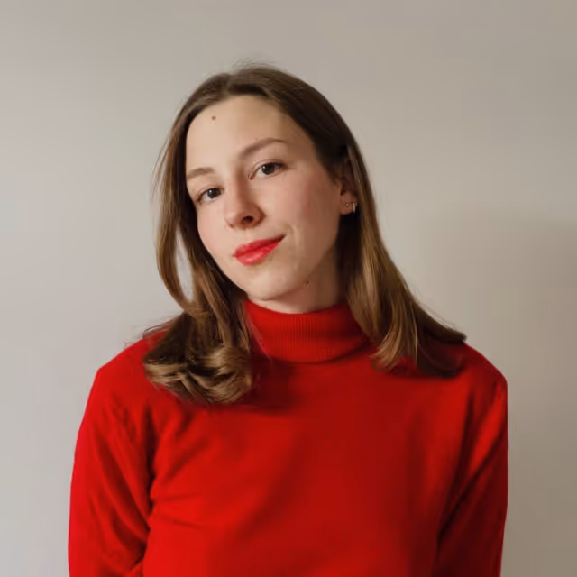 Young woman with shoulder-length brown hair wearing a red turtleneck sweater against a plain beige background.