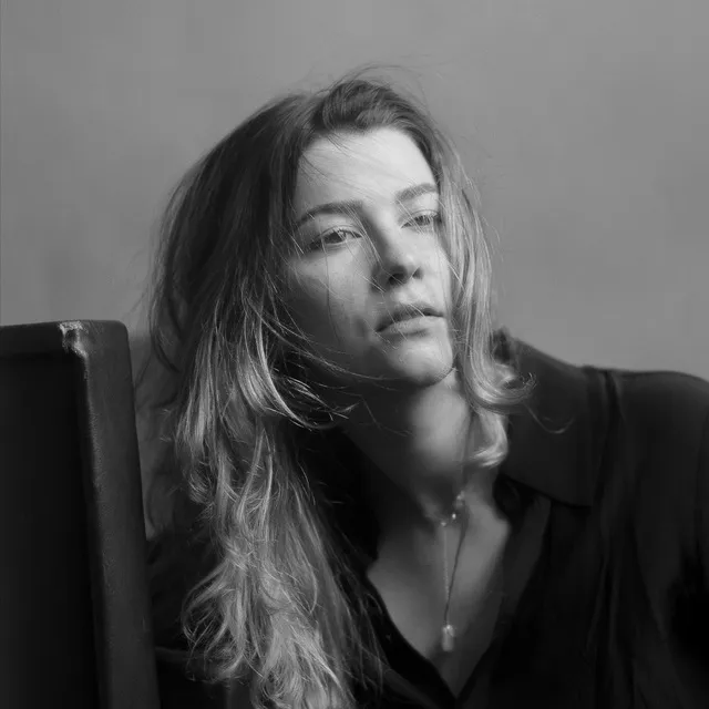 Black and white portrait of a woman with long hair partially covering her face, wearing a dark coat, standing outdoors.