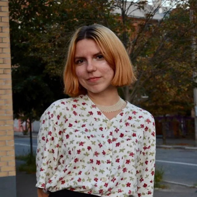 Young woman with long brown hair and glasses taking a selfie outdoors near glass doors.