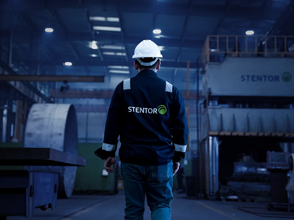 Worker wearing a white hard hat and STENTOR jacket walking inside a dimly lit industrial facility.