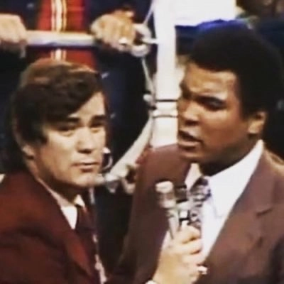 Bob Sheridan holds a microphone while interviewing Muhammad Ali beside a boxing ring.