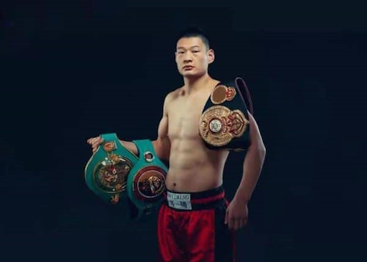 Zhaoxin Zhang in red trunks holding championship belts against a dark background.