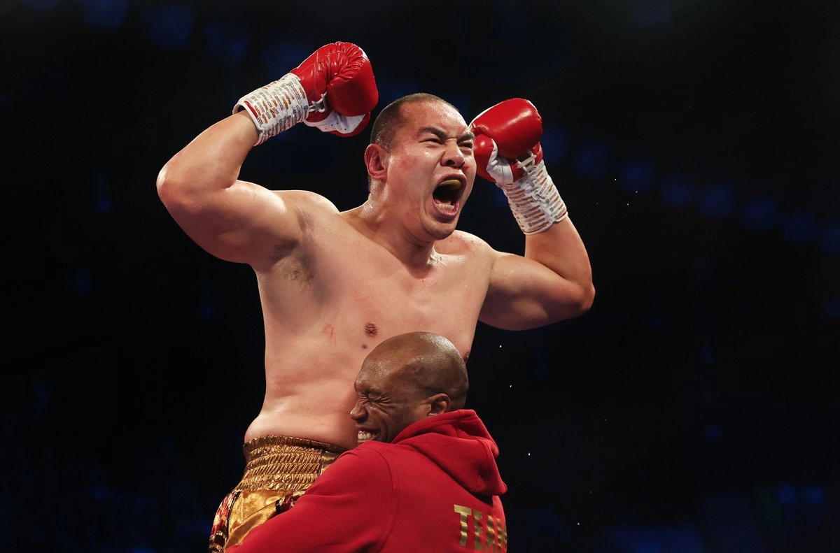 Zhilei Zhang raising both red boxing gloves as a man in a red top embraces him.