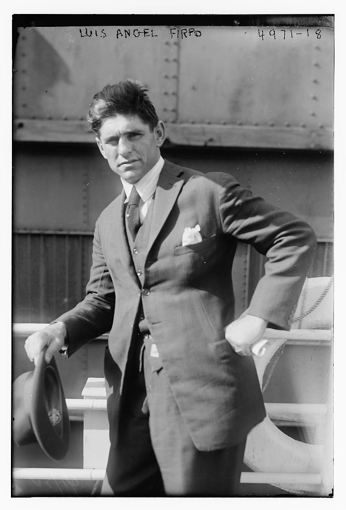 Black-and-white portrait of Luis Angel Firpo in a suit and tie, holding a hat beside ring ropes.
