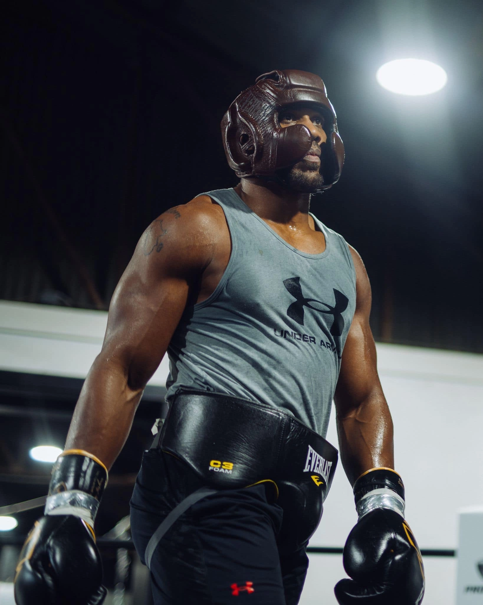 Anthony Joshua wearing brown headgear, black gloves and a grey training vest in a boxing ring.