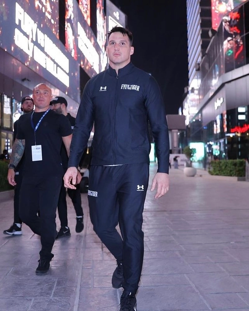 Simon Kean in a dark tracksuit walking along an illuminated pedestrian street with men behind him.