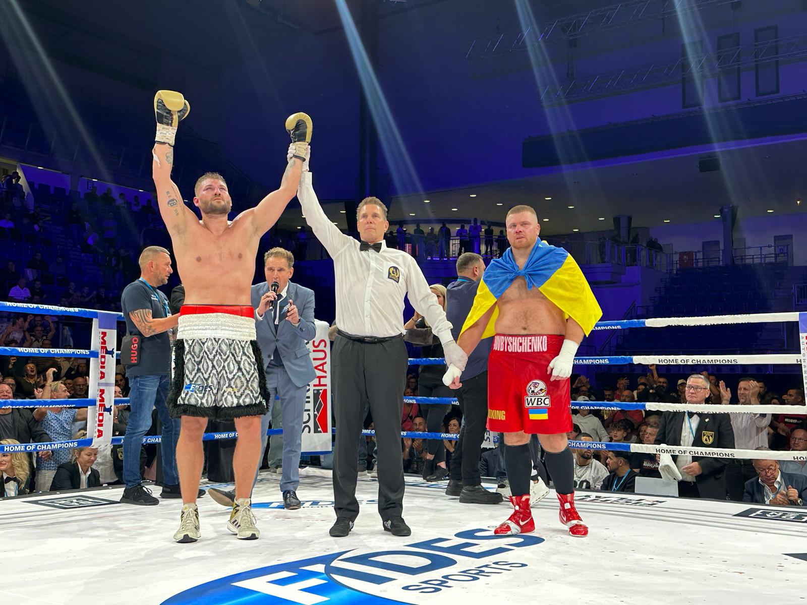 Tom Schwarz with both gloves raised as the referee lifts his arm, beside Kostiantyn Dovbyshchenko draped in a Ukrainian flag.