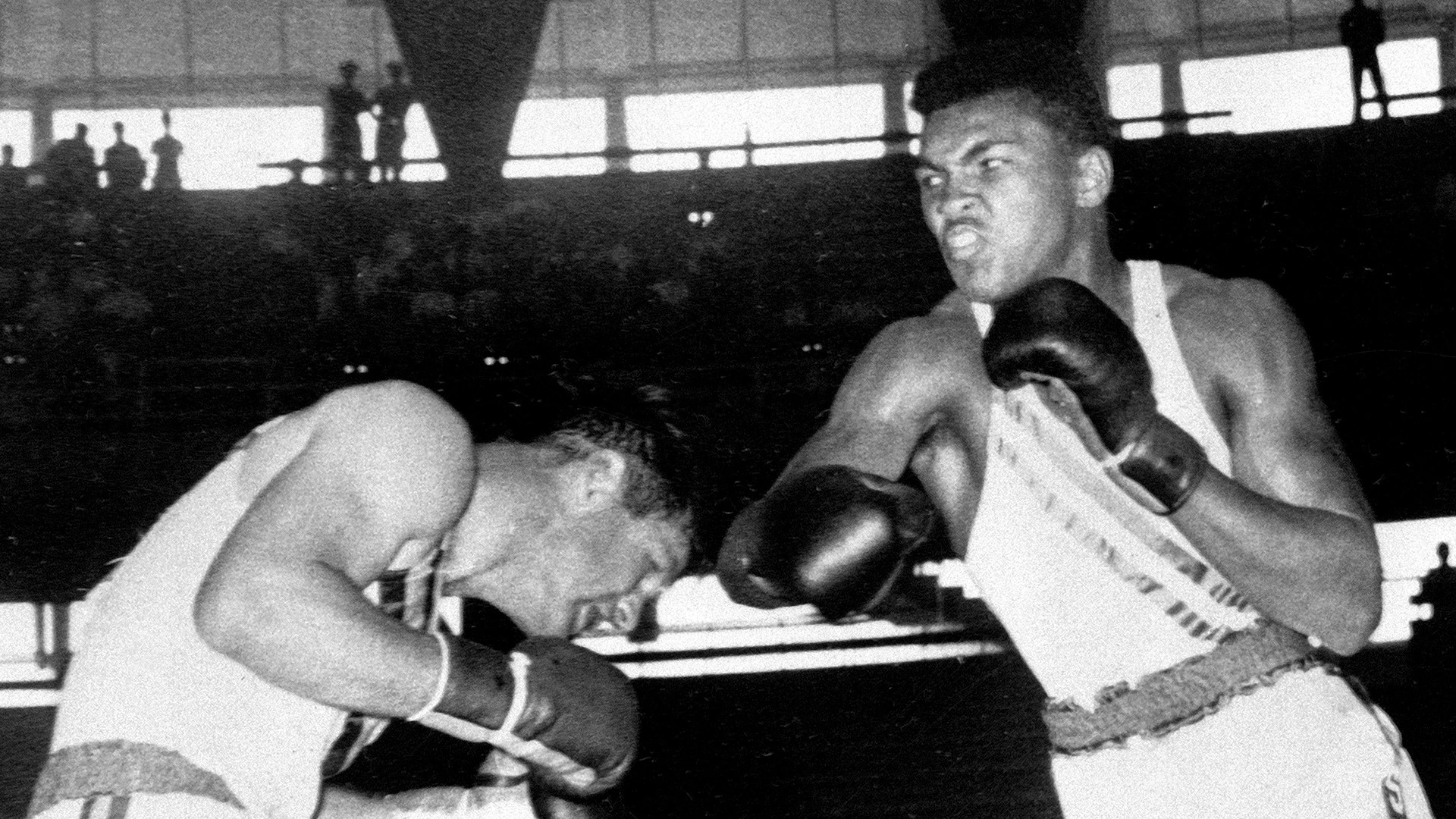 Black-and-white photograph of Muhammad Ali in a white boxing vest and dark gloves facing an opponent bending forward.