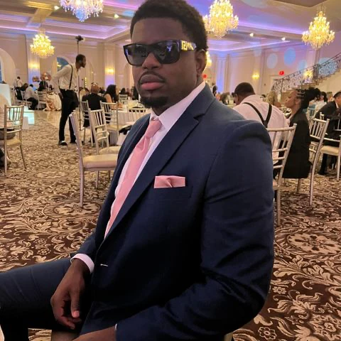Cassius Chaney seated at a formal event wearing a dark suit, pink tie and sunglasses.
