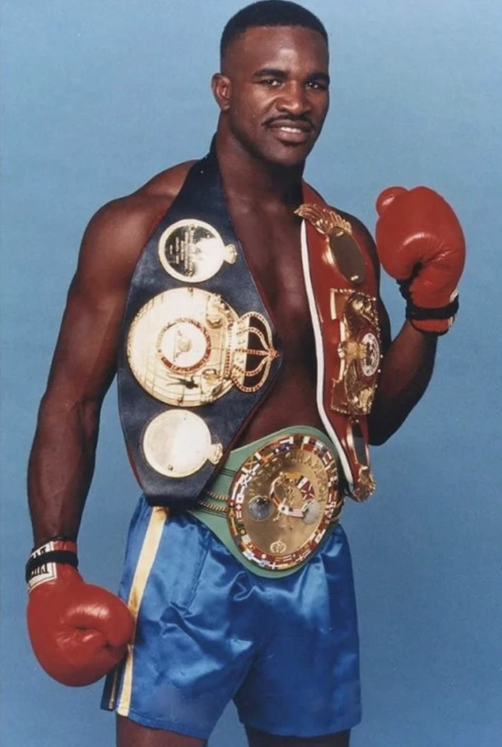 Evander Holyfield poses in red boxing gloves with three championship belts.