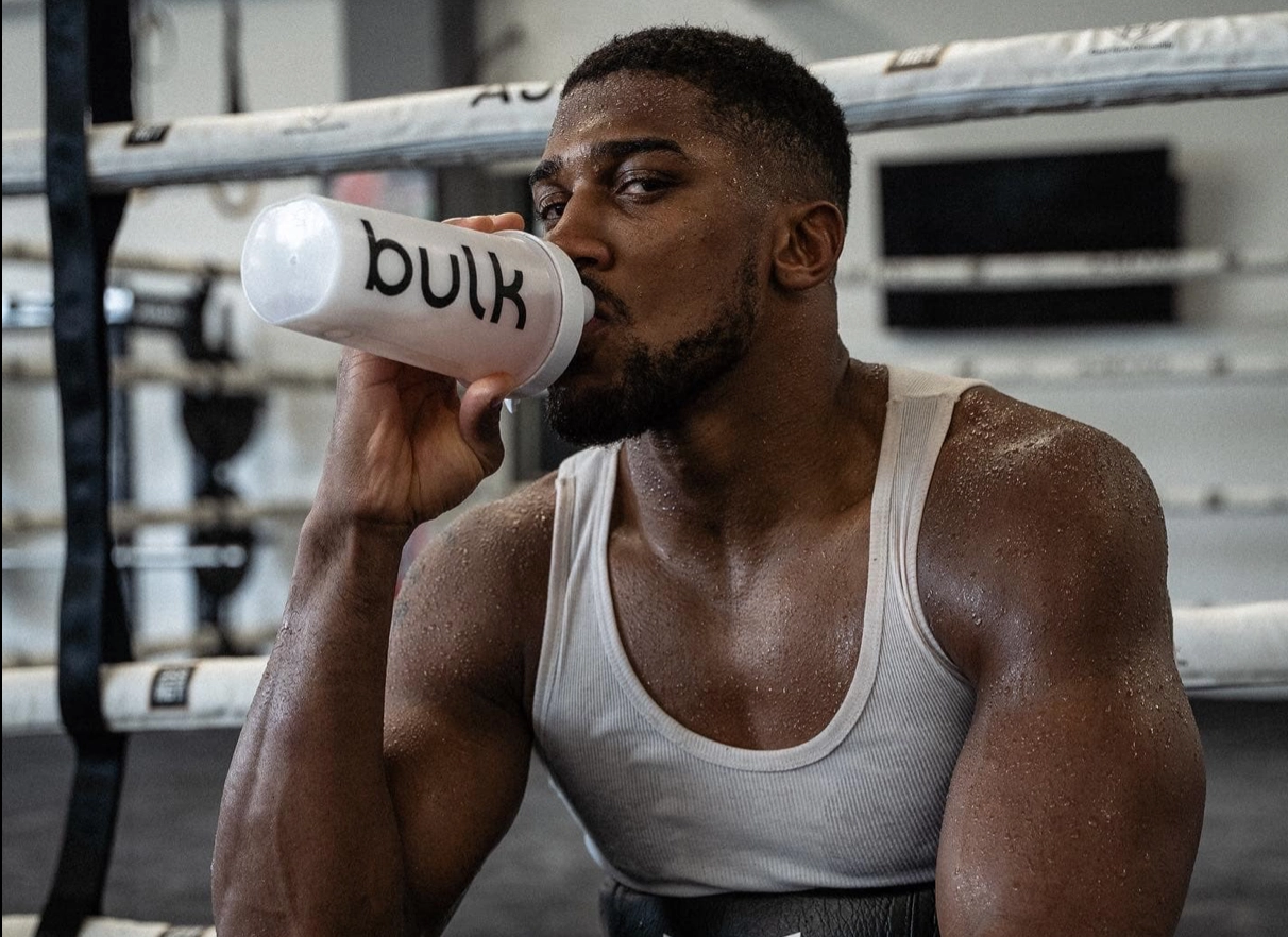 Anthony Joshua drinks from a white bottle beside the ropes in a boxing gym.