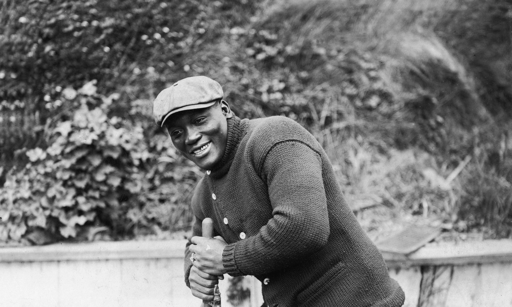 Black-and-white photograph of Jack Johnson smiling outdoors in a flat cap and knitted cardigan.