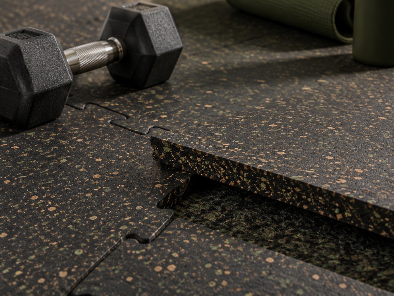 Close-up of interlocking cork-rubber gym tiles with one tile lifted to show its thickness and the underlayment beneath, a hex dumbbell beside it