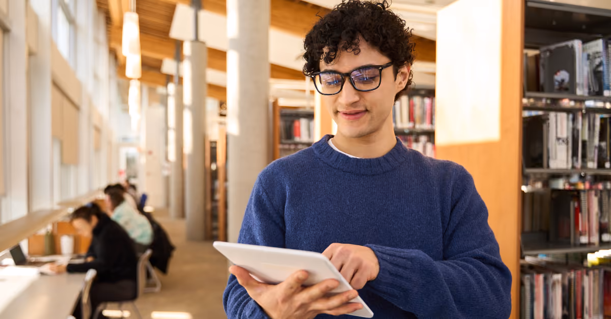 Young man in glasses using a tablet in a bright library with shelves and people studying.
