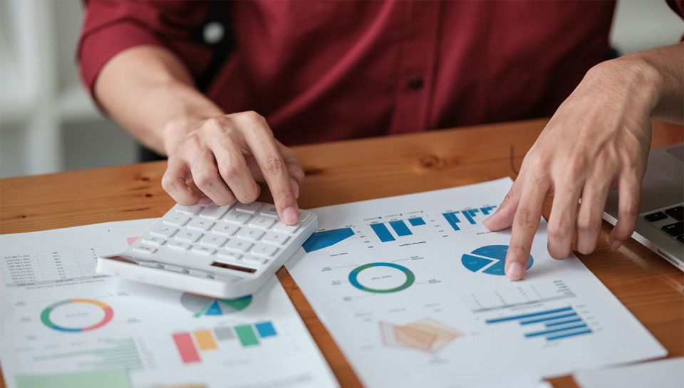 Person using calculator and pointing at charts and graphs on printed papers on a wooden table.