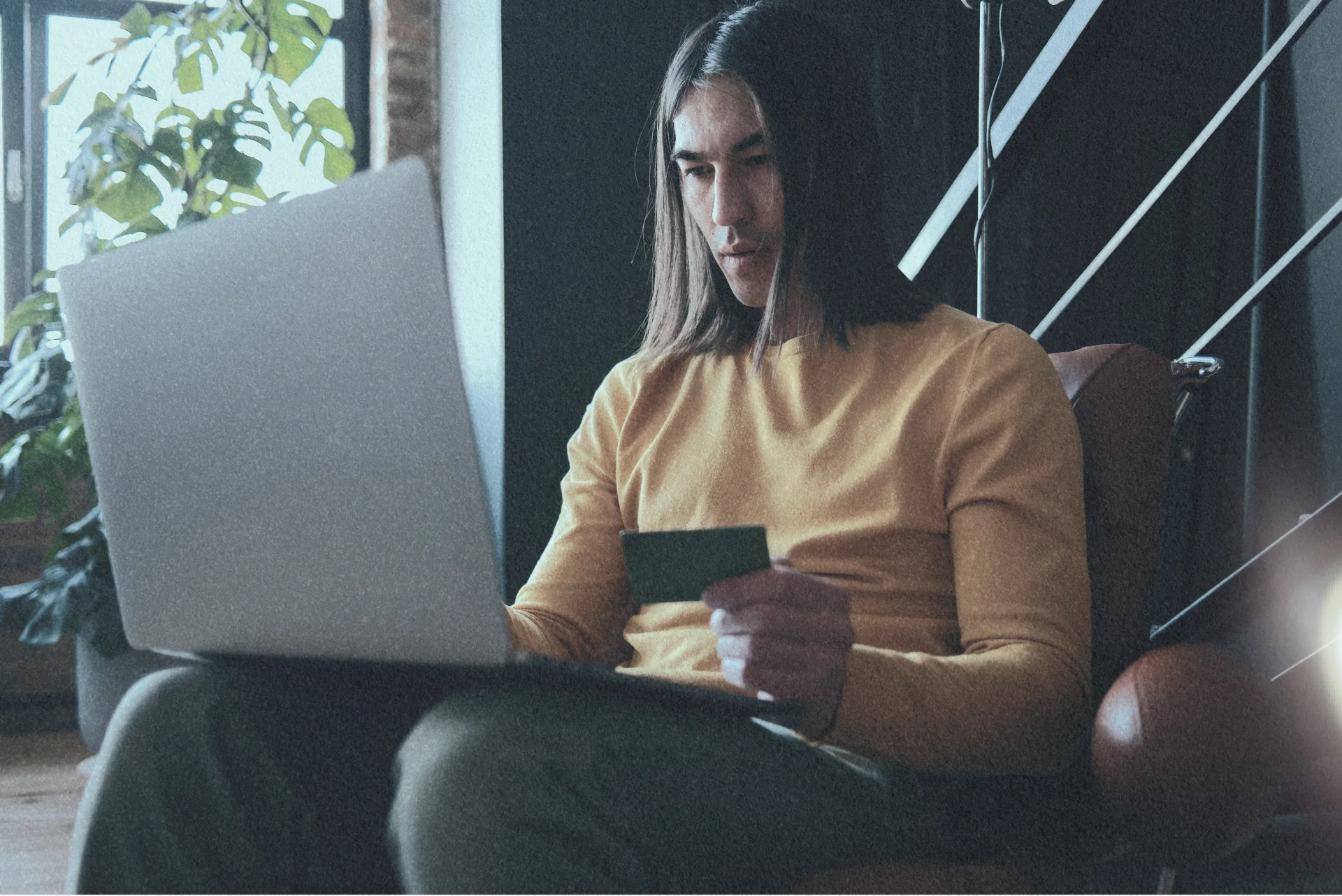 Person with long hair wearing a yellow sweater sitting in a chair using a laptop and holding a credit card indoors.