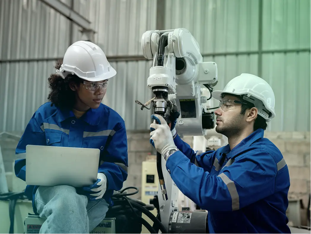 Two engineers in blue workwear, hard hats, and safety glasses inspect an industrial robot; one uses a laptop as the other adjusts its arm.