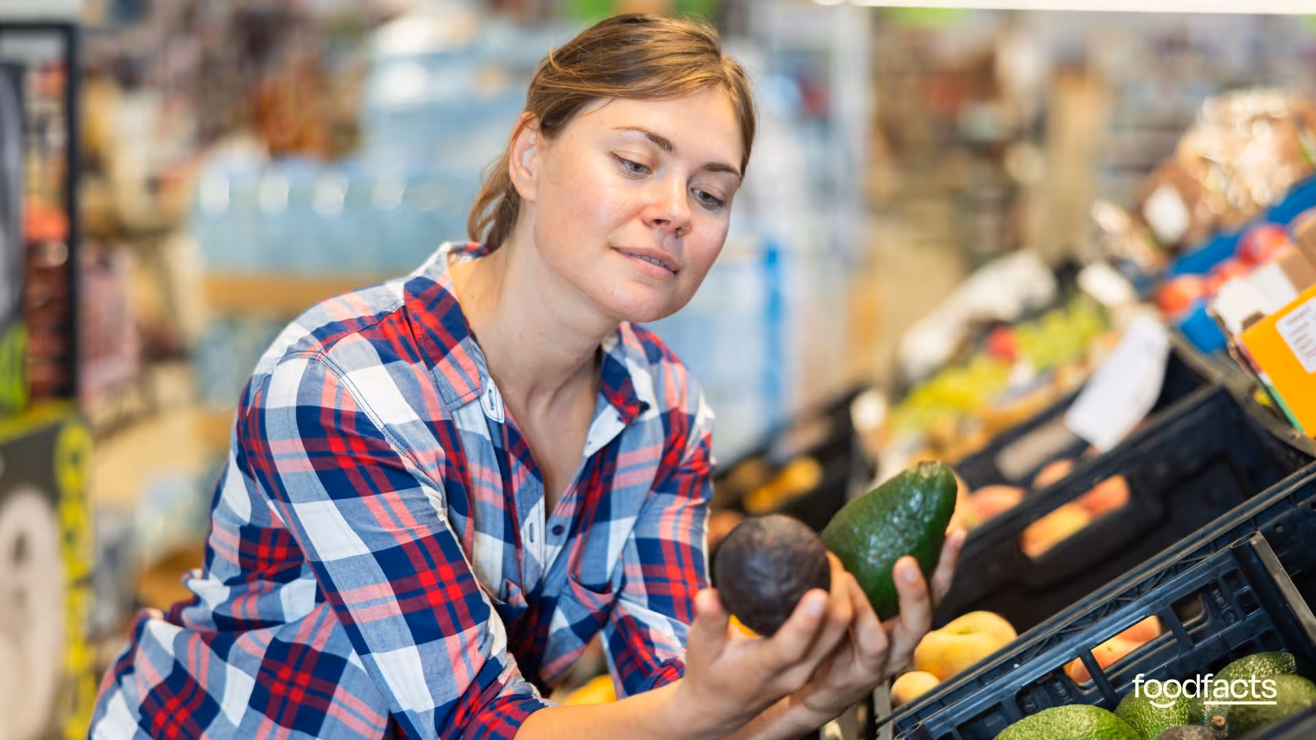 A woman in a plaid shirt closely inspects two avocados while shopping in the produce section of a grocery store. The image, featured on FoodFacts.org, relates to the debate over the necessity of best-before dates, highlighting consumer decision-making and food waste reduction through personal judgment and visual inspection of freshness.
