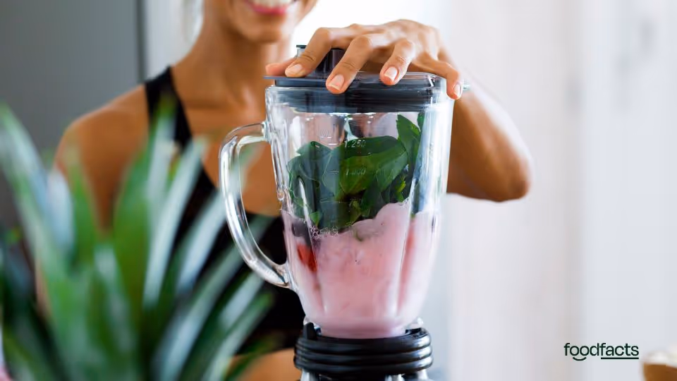 A woman smiles as she prepares to blend a smoothie containing leafy greens and pink fruit yogurt in a blender. The image accompanies an article investigating Jessie Inchauspé’s advice to avoid smoothies for blood sugar management. It visually contrasts the popular image of smoothies as healthy with FoodFacts.org’s scientific analysis of how different ingredients and preparation methods affect glucose response.