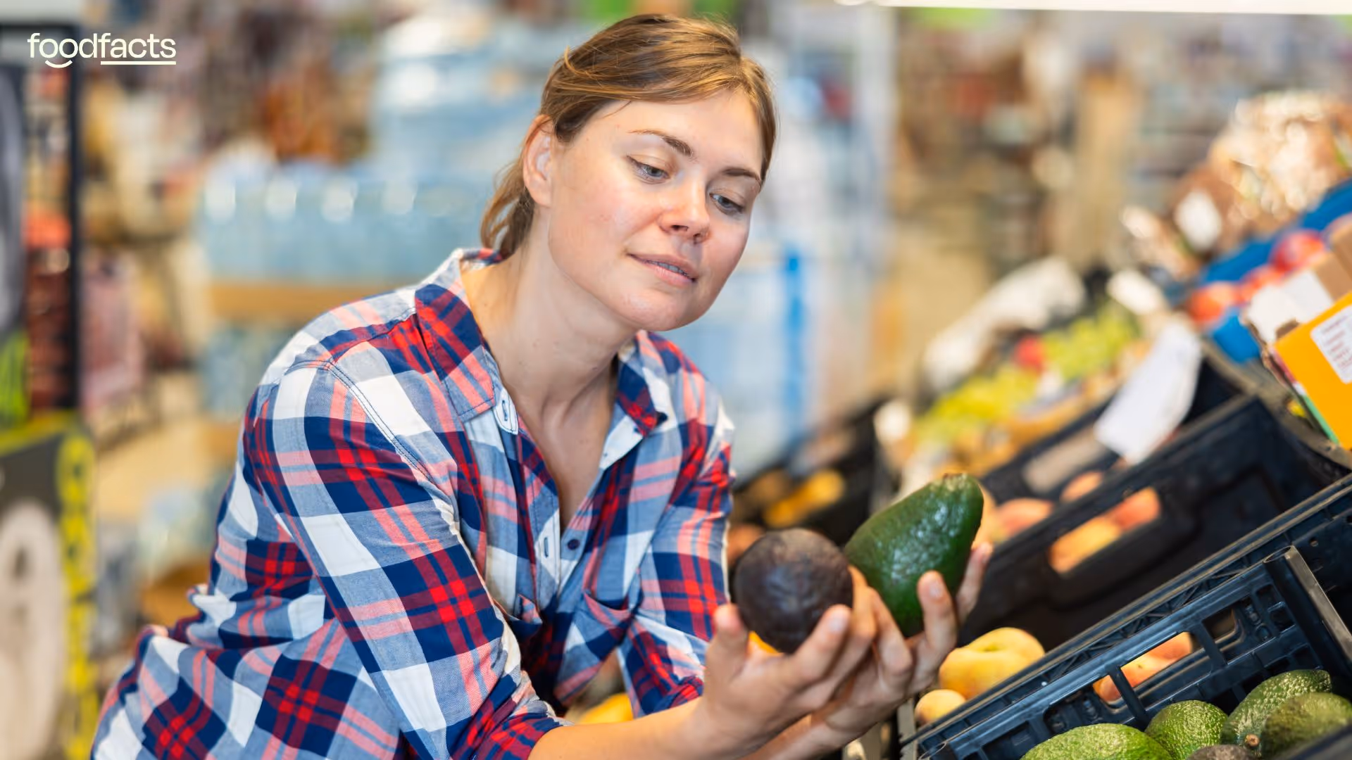 A woman in a plaid shirt closely inspects two avocados while shopping in the produce section of a grocery store. The image, featured on FoodFacts.org, relates to the debate over the necessity of best-before dates, highlighting consumer decision-making and food waste reduction through personal judgment and visual inspection of freshness.