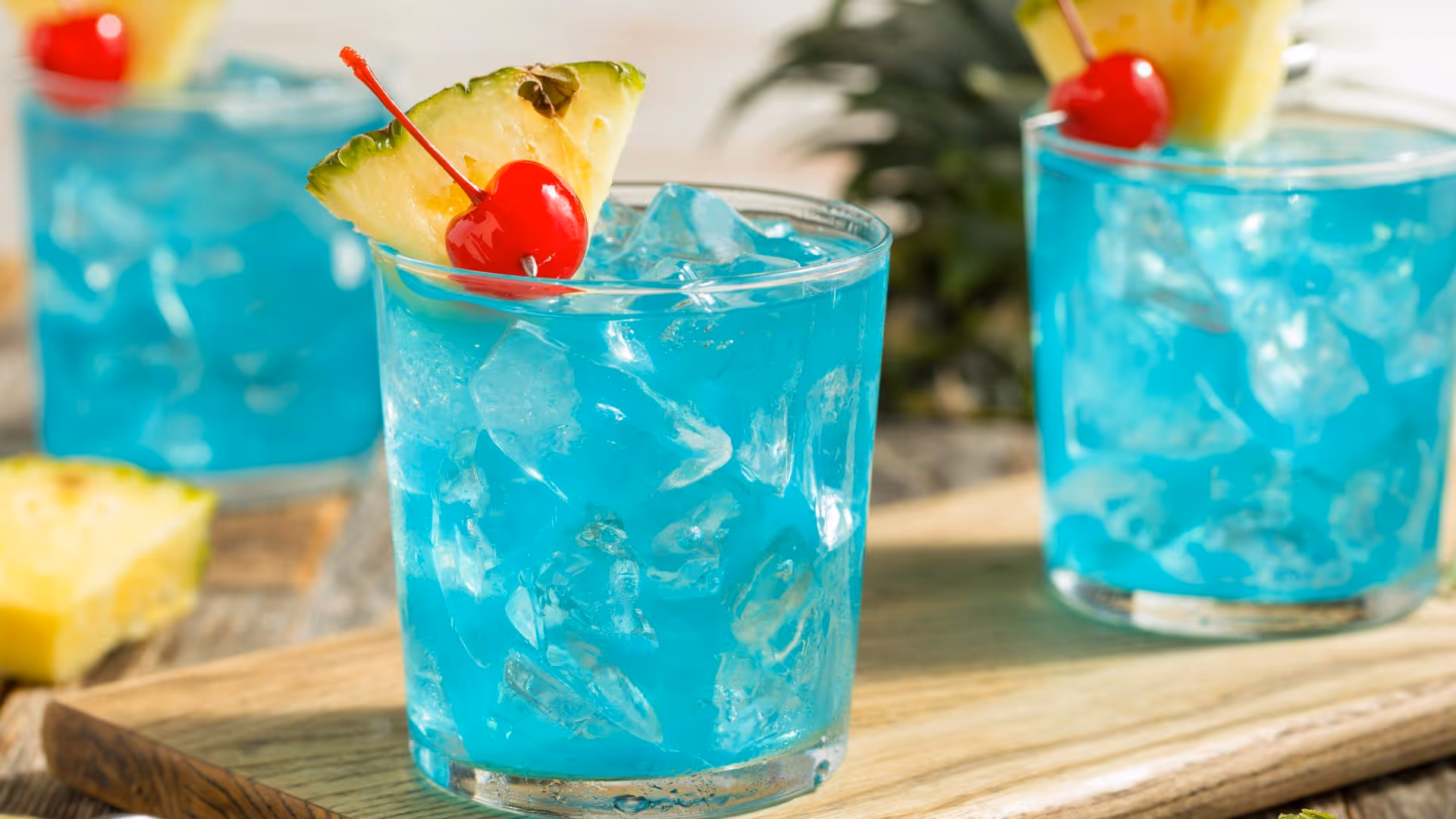 Three glasses filled with blue liquids are adorned with a lice of pineapple and a cocktail cherry.