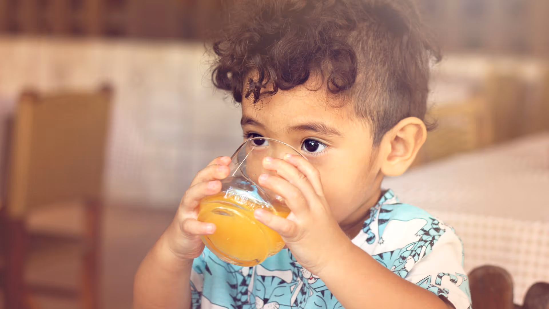 A young child holds and drinks a glass of orange juice with both hands.