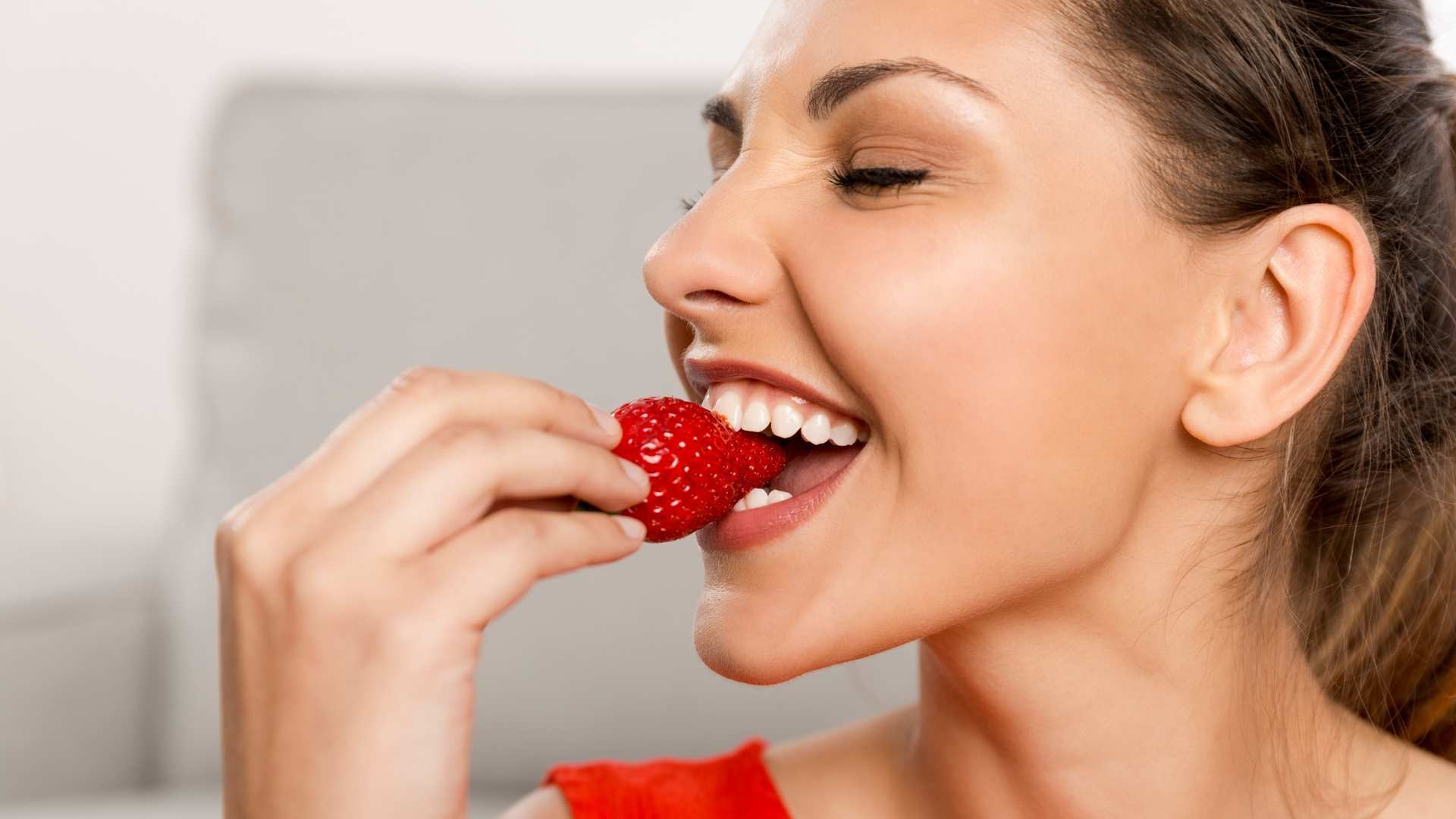 A happy woman holds a strawberry in her hand whilst taking a bite of it