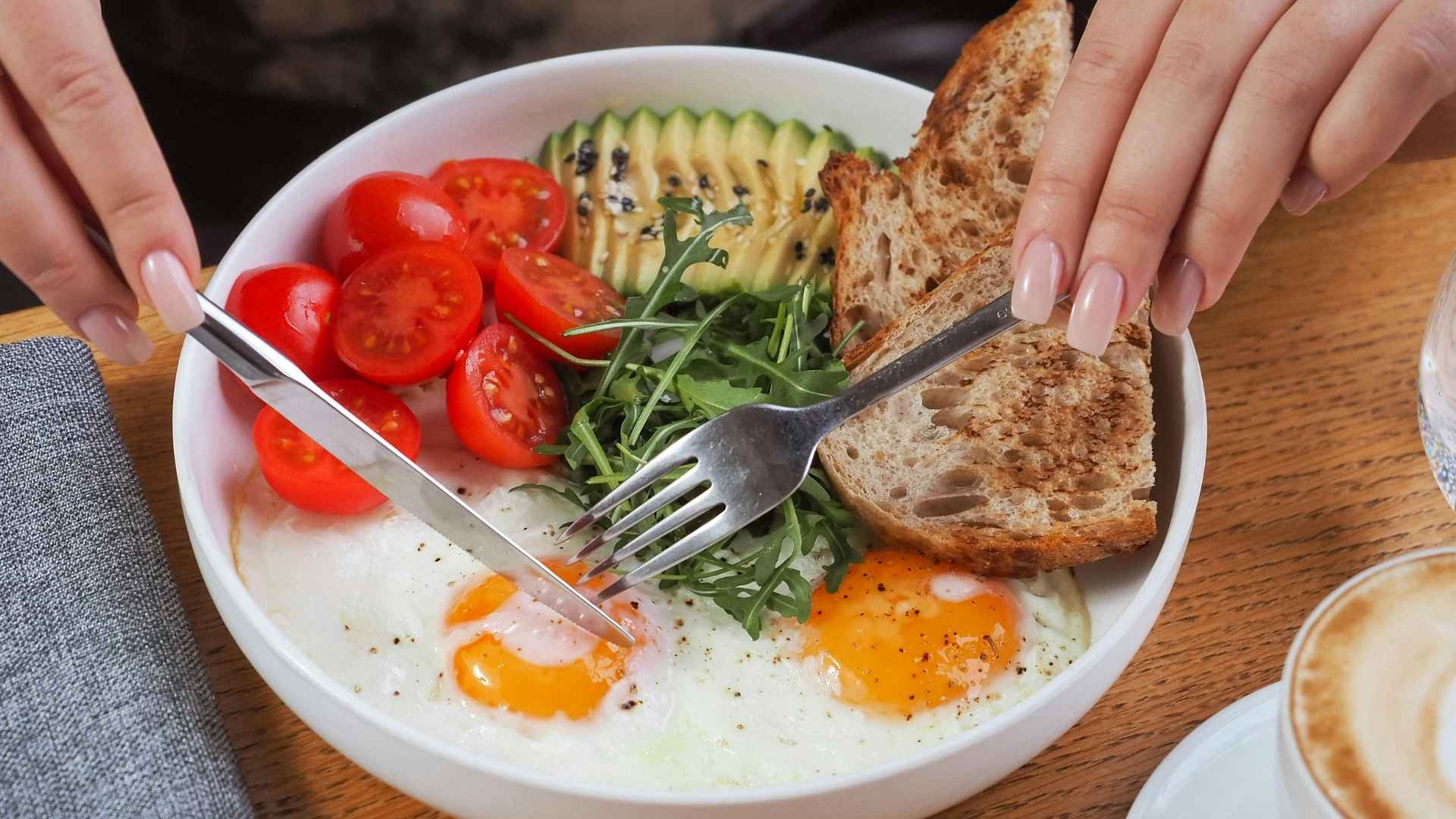 A bowl of food containing eggs, tomatoes, bread, avocado, and fresh greens
