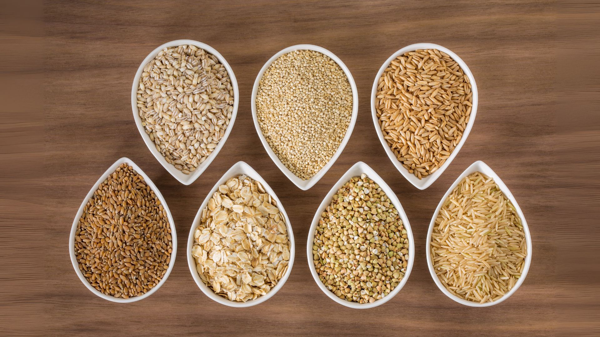 Seven white bowls are seen, each containing a different type of whole grain, such as oats, barley, and rice