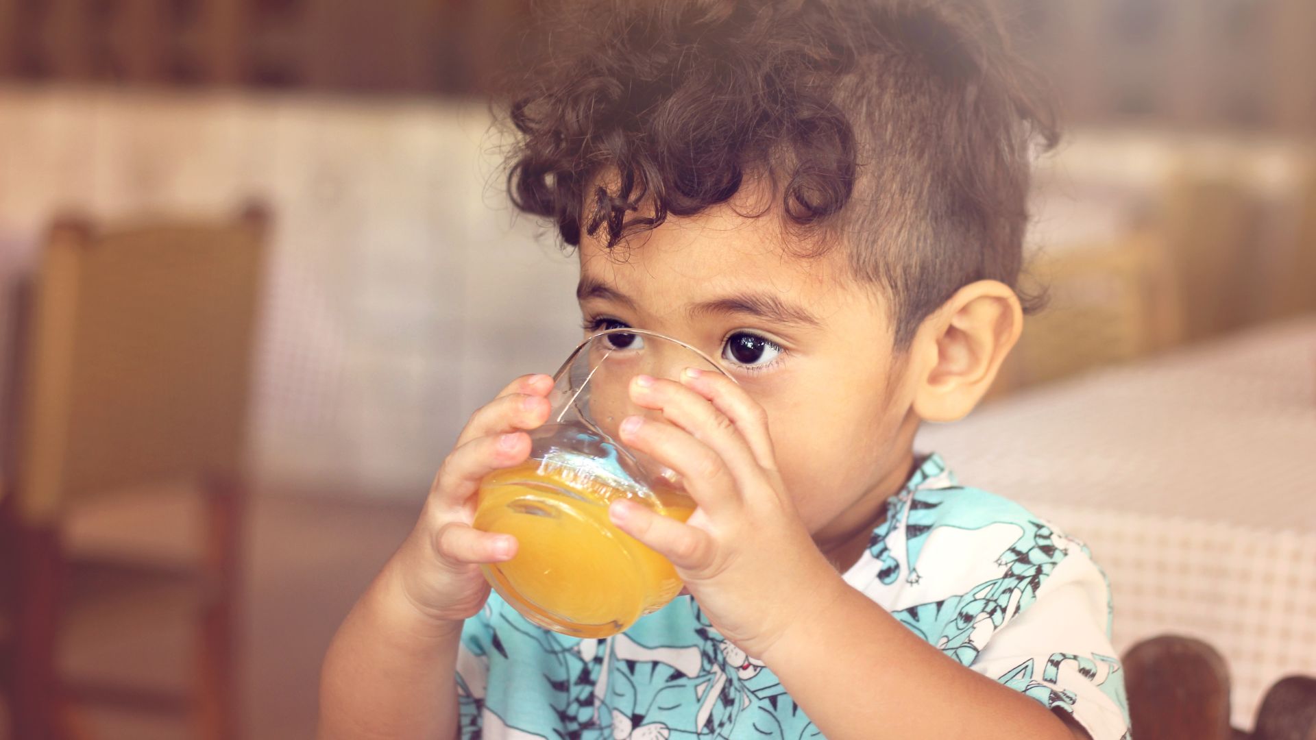 A young child holds and drinks a glass of orange juice with both hands.