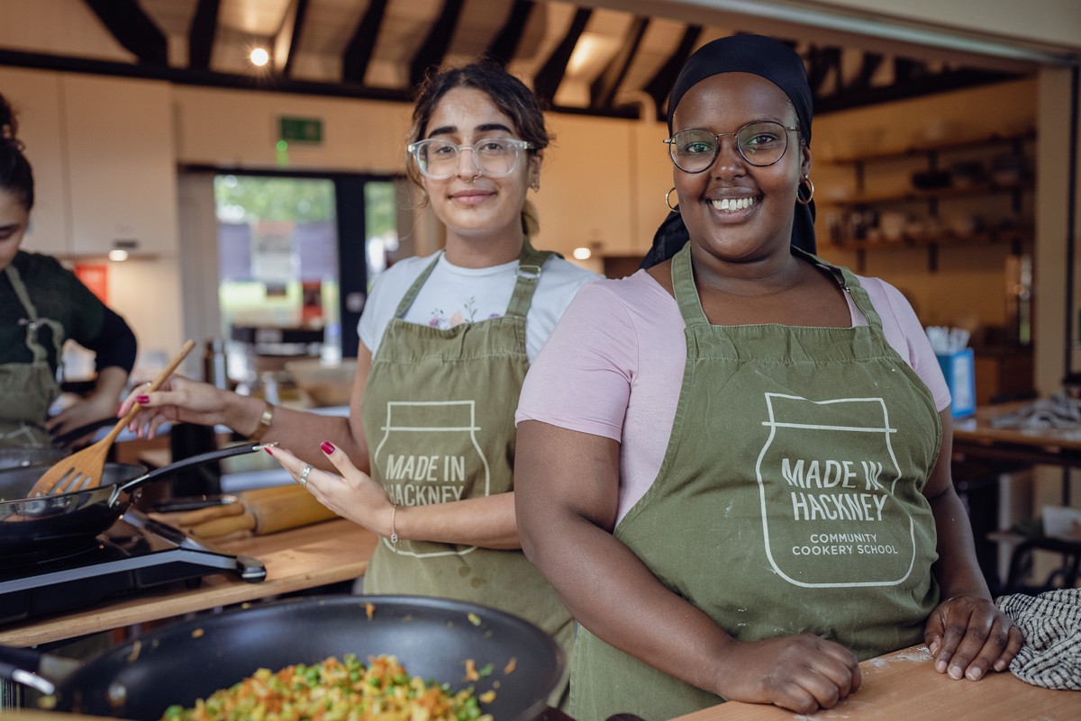 Two young women look at the camera in a kitchen enjoying lessons on cooking plant-based meals in a community kitchen in London, known as Made in Hackney