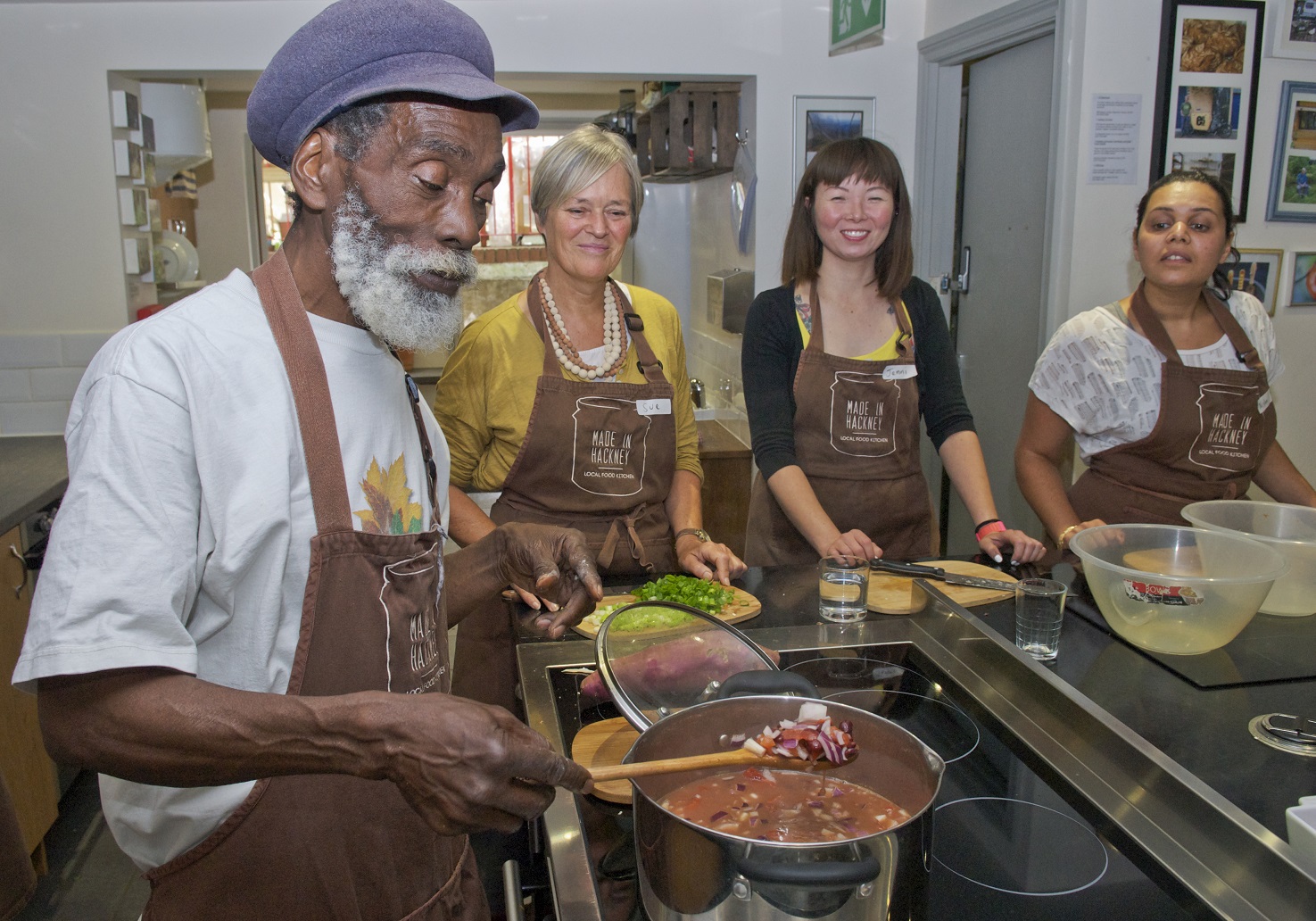 A collection of people gather around a cooking pot in a kitchen enjoying lessons on cooking plant-based meals in a community kitchen in London, known as Made in Hackney