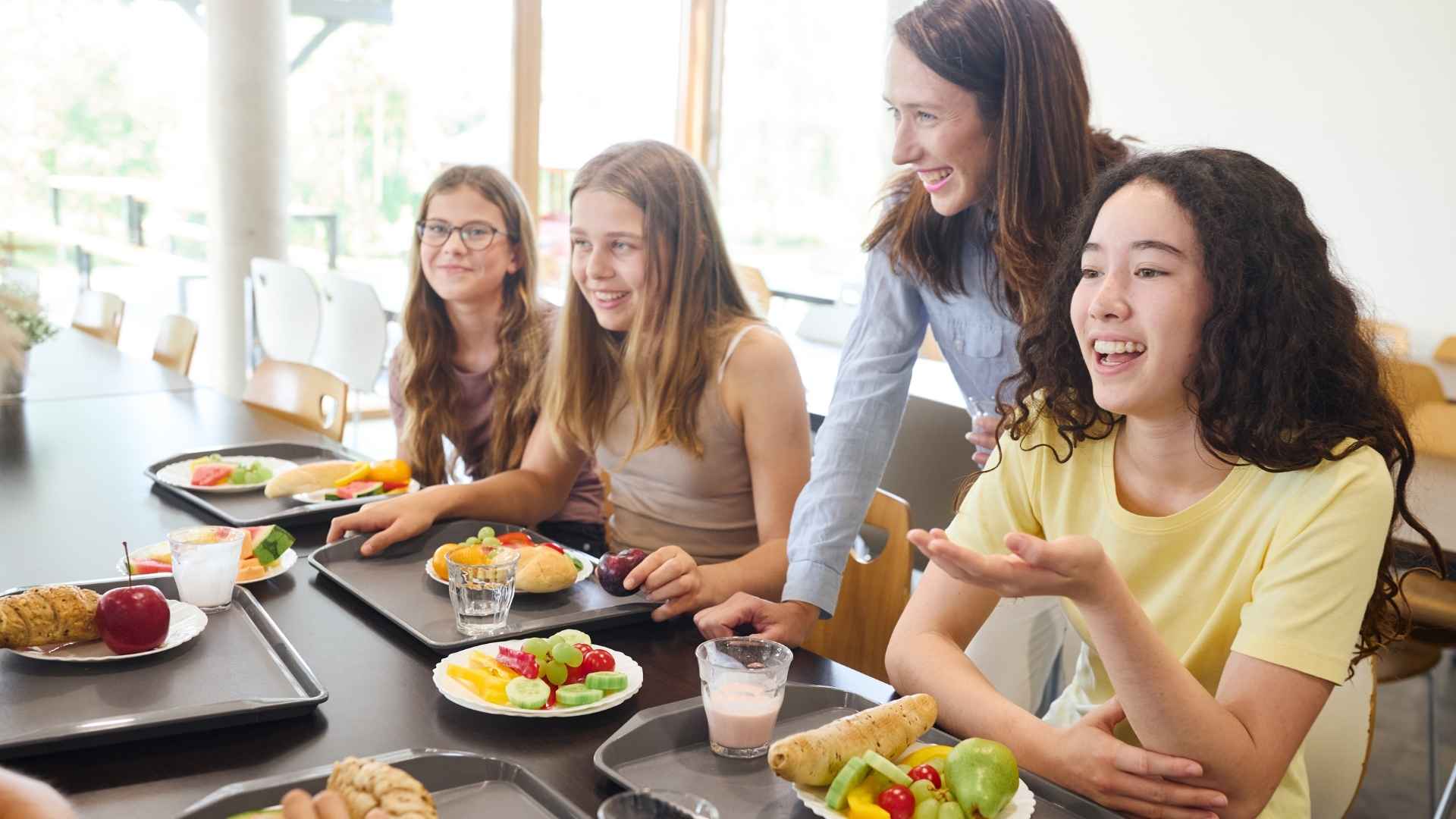 A group of high school children sit around a lunch table with trays of healthy food