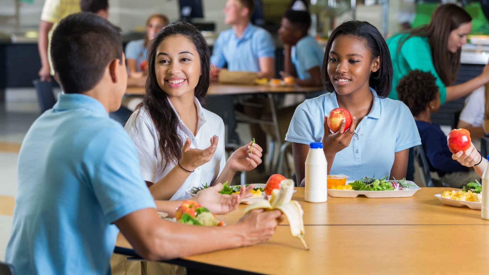 Older school children sit at a cafeteria table eating a school meal together