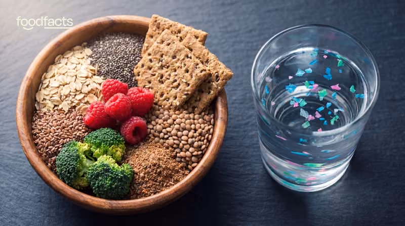 Bowl of high-fibre foods including rolled oats, chia seeds, raspberries, broccoli and crackers next to a glass of water containing visible coloured microplastic particles