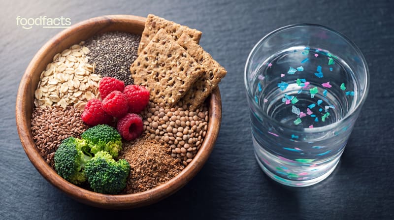 Bowl of high-fibre foods including rolled oats, chia seeds, raspberries, broccoli and crackers next to a glass of water containing visible coloured microplastic particles