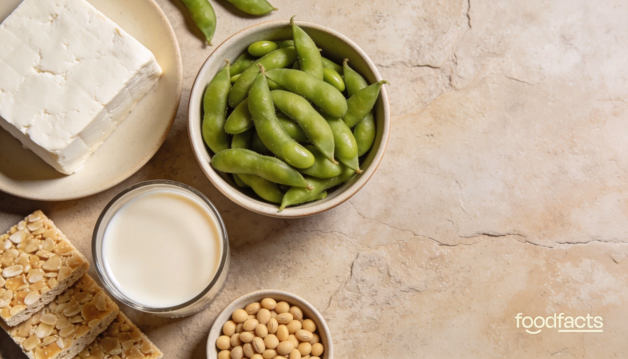 An overhead spread of whole soy foods — a block of firm tofu, a bowl of edamame, a glass of soy milk and dried soybeans